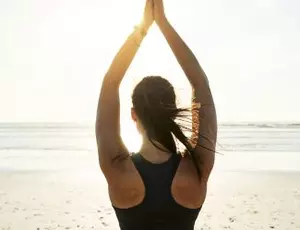 A woman stands on a beach with her hands above her head