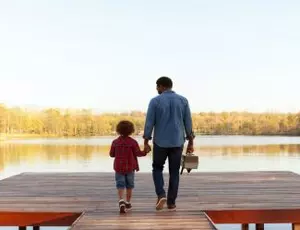 A man and his son walk onto a dock on a lake with a fishing tackle box