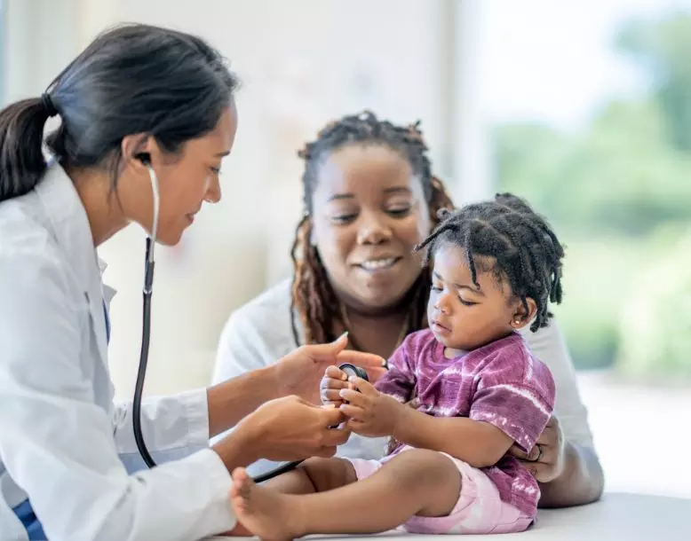 A young child plays with a doctor's stethoscope as the mother watches smiling